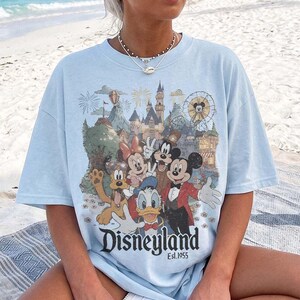 May include: A light blue t-shirt featuring a graphic print of Mickey Mouse, Minnie Mouse, Donald Duck, Goofy, and Pluto in front of a castle and ferris wheel. The text Disneyland Est. 1955 is printed in black below the characters on the front.