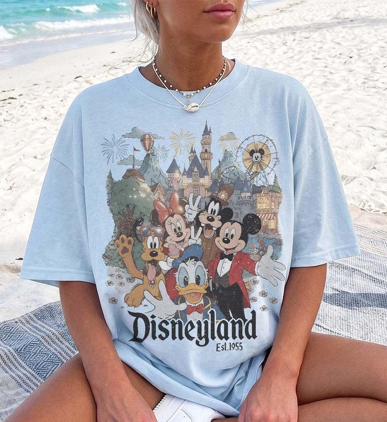 May include: A light blue t-shirt featuring a graphic print of Mickey Mouse, Minnie Mouse, Donald Duck, Goofy, and Pluto in front of a castle and ferris wheel. The text Disneyland Est. 1955 is printed in black below the characters on the front.