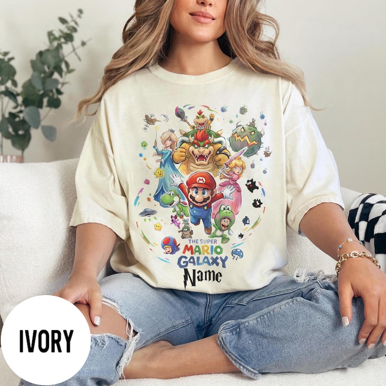 Puede incluir: Camiseta de color marfil con un gr&aacute;fico colorido de Mario y otros personajes del juego Super Mario Galaxy. El texto "THE SUPER MARIO GALAXY Name" est&aacute; impreso debajo. La camiseta tiene un corte holgado.