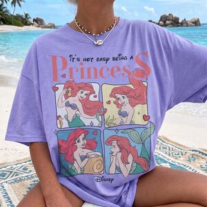 Puede incluir: Una camiseta de manga corta de color morado claro con un estampado gr&aacute;fico del personaje Ariel de La Sirenita en cuatro paneles. El texto dice It's not easy being a Princess y Disney. La camiseta tiene cuello redondo y un corte holgado.