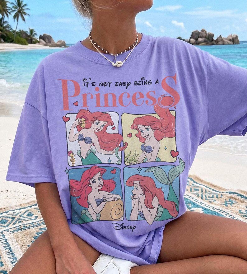 Puede incluir: Una camiseta de manga corta de color morado claro con un estampado gr&aacute;fico del personaje Ariel de La Sirenita en cuatro paneles. El texto dice It's not easy being a Princess y Disney. La camiseta tiene cuello redondo y un corte holgado.