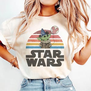 May include: Cream-colored t-shirt featuring a graphic of Grogu from Star Wars holding a Mickey Mouse balloon, set against a retro sunset design. The Star Wars logo is printed below the graphic.