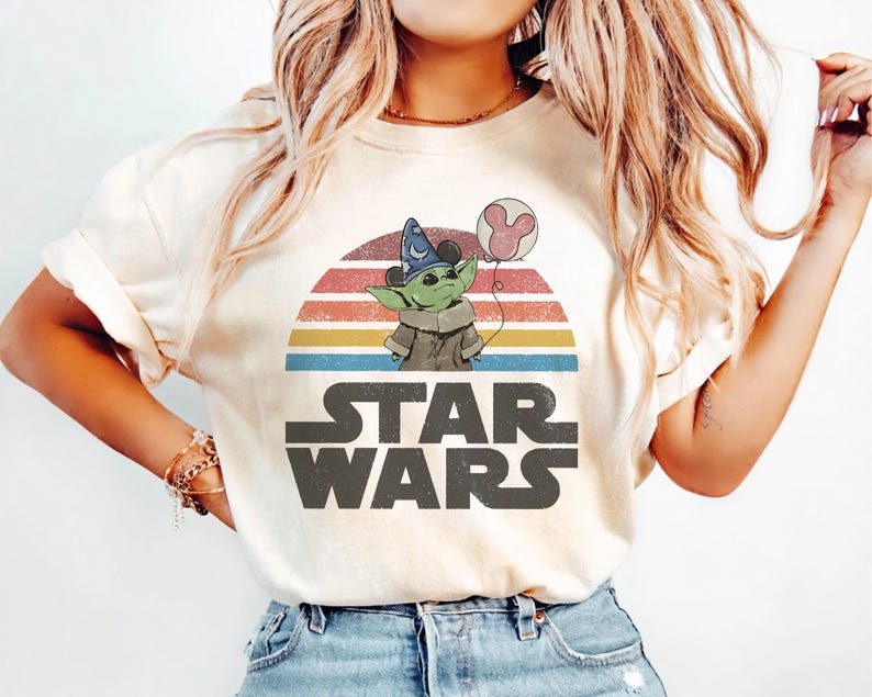 May include: Cream-colored t-shirt featuring a graphic of Grogu from Star Wars holding a Mickey Mouse balloon, set against a retro sunset design. The Star Wars logo is printed below the graphic.