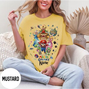 Puede incluir: Camiseta amarilla mostaza con un gr&aacute;fico colorido de Mario y otros personajes del juego Super Mario Galaxy. El texto "THE SUPER MARIO GALAXY Name" est&aacute; impreso debajo. La camiseta es de estilo casual y de manga corta.