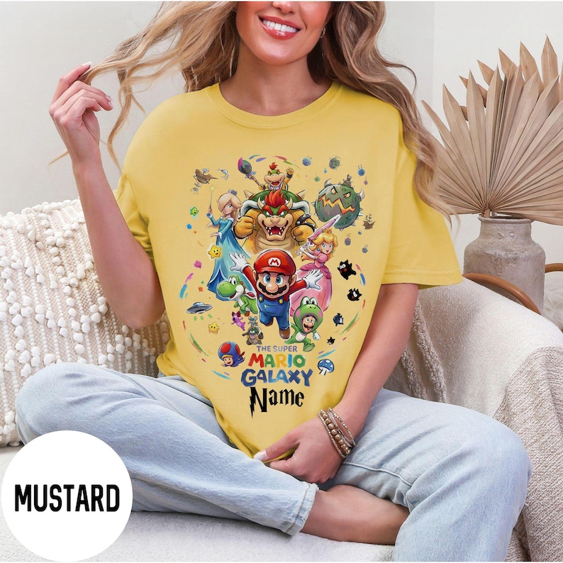 Puede incluir: Camiseta amarilla mostaza con un gr&aacute;fico colorido de Mario y otros personajes del juego Super Mario Galaxy. El texto "THE SUPER MARIO GALAXY Name" est&aacute; impreso debajo. La camiseta es de estilo casual y de manga corta.