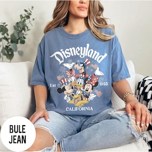 May include: Blue t-shirt with a Disneyland graphic featuring Mickey Mouse, Minnie Mouse, Donald Duck, Goofy, and Pluto in patriotic attire. The shirt has the words "Disneyland", "Est 1955", and "California" printed on it.
