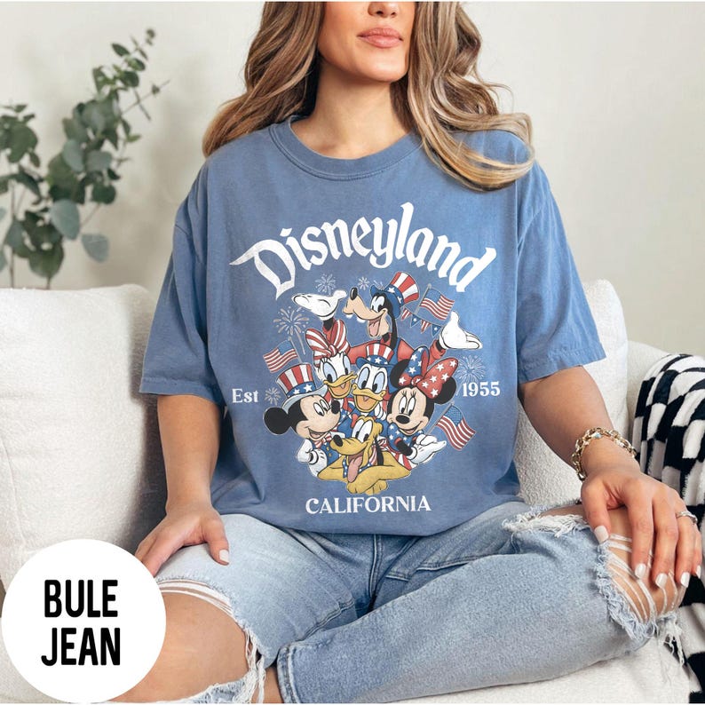 May include: Blue t-shirt with a Disneyland graphic featuring Mickey Mouse, Minnie Mouse, Donald Duck, Goofy, and Pluto in patriotic attire. The shirt has the words "Disneyland", "Est 1955", and "California" printed on it.