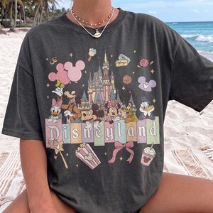 May include: A charcoal grey t-shirt featuring a colourful graphic print of the Disneyland castle, Mickey Mouse, Minnie Mouse, Donald Duck, Daisy Duck, Goofy, and Pluto. The design includes pastel-coloured rectangular blocks with the text Disneyland.
