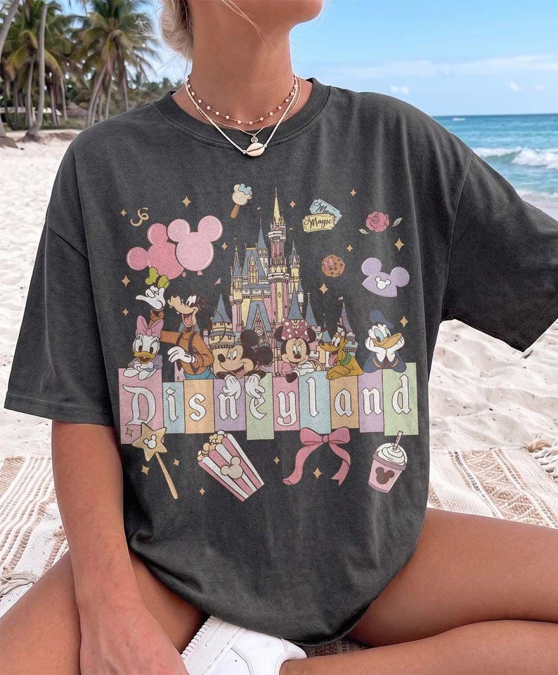 May include: A charcoal grey t-shirt featuring a colourful graphic print of the Disneyland castle, Mickey Mouse, Minnie Mouse, Donald Duck, Daisy Duck, Goofy, and Pluto. The design includes pastel-coloured rectangular blocks with the text Disneyland.
