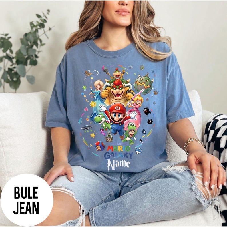 Puede incluir: Camiseta azul claro con un dise&ntilde;o gr&aacute;fico colorido de Mario y otros personajes de Super Mario Galaxy. El texto "MARIO GALAXY NAME" est&aacute; impreso debajo. La camiseta est&aacute; hecha de un material suave.