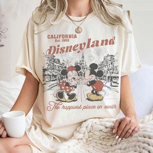 May include: Cream-colored t-shirt featuring a Disneyland graphic. The design includes Mickey and Minnie Mouse, the words "California Disneyland Est. 1955," and "The happiest place on earth." The graphic is in shades of brown and red.