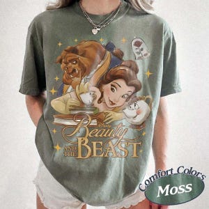 May include: A moss green short-sleeve t-shirt featuring a graphic print of Belle, the Beast, Chip, and Mrs. Potts from Beauty and the Beast. The design includes a rose in a glass dome, yellow stars, and the text Beauty and the Beast in gold script.
