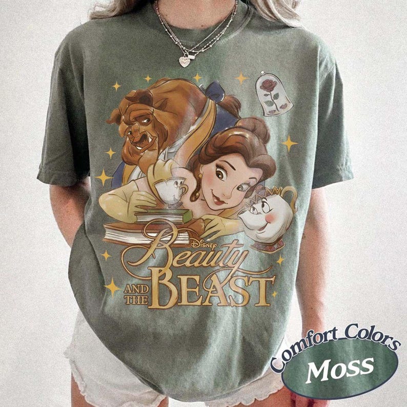 May include: A moss green short-sleeve t-shirt featuring a graphic print of Belle, the Beast, Chip, and Mrs. Potts from Beauty and the Beast. The design includes a rose in a glass dome, yellow stars, and the text Beauty and the Beast in gold script.