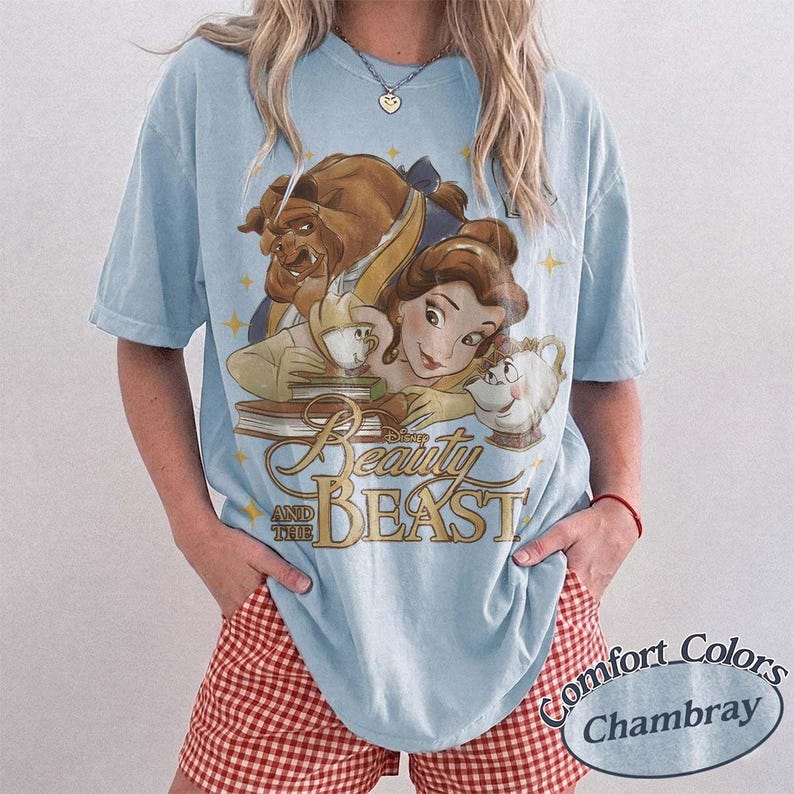 May include: A light blue short-sleeved t-shirt featuring a graphic print of Belle, the Beast, and Mrs. Potts from Beauty and the Beast. The design includes gold-coloured text that reads Beauty and the Beast, surrounded by small yellow star accents.