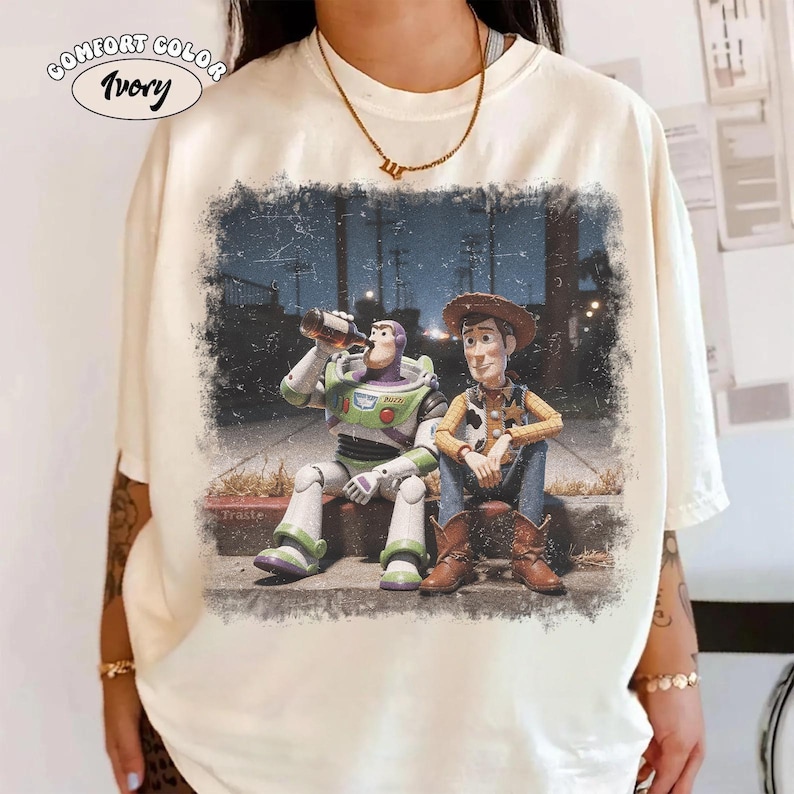 May include: An ivory t-shirt featuring a graphic print of Buzz Lightyear and Woody sitting together outdoors at night. Buzz Lightyear holds a brown bottle, while Woody sits beside him. The graphic has a distressed, vintage-style texture.
