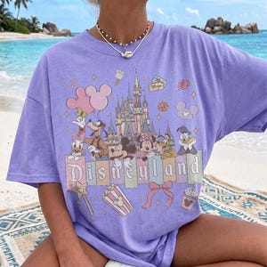 May include: A light purple short-sleeve t-shirt featuring a graphic print of a castle, Mickey Mouse, Minnie Mouse, Goofy, Donald Duck, Daisy Duck, and Pluto. The text Disneyland is printed in the centre with icons of balloons, popcorn, and a bow.
