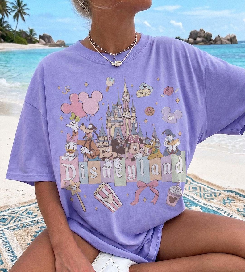 May include: A light purple short-sleeve t-shirt featuring a graphic print of a castle, Mickey Mouse, Minnie Mouse, Goofy, Donald Duck, Daisy Duck, and Pluto. The text Disneyland is printed in the centre with icons of balloons, popcorn, and a bow.