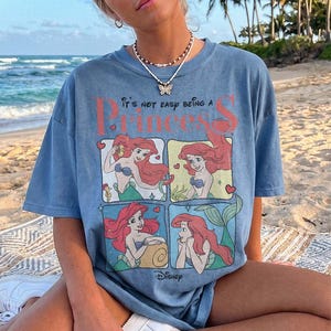 Puede incluir: Una camiseta azul de manga corta con un estampado gr&aacute;fico del personaje Ariel de La Sirenita en cuatro paneles distintos. El texto dice It's not easy being a Princess. El dise&ntilde;o incluye corazones rojos y el logotipo de Disney.