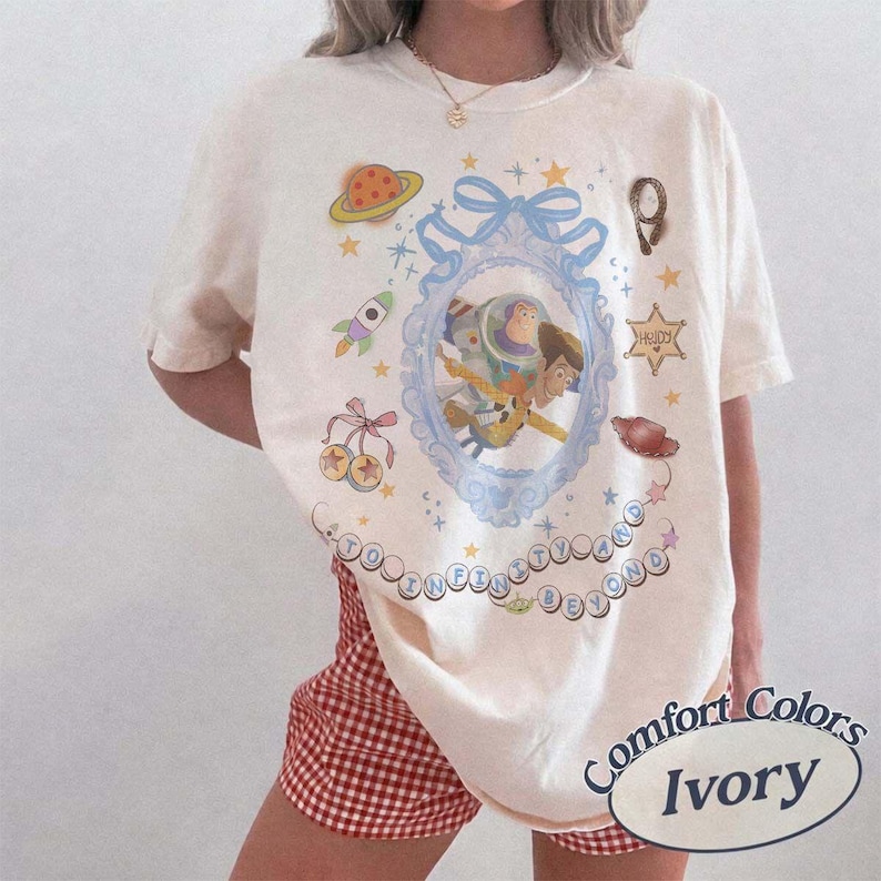 Peut inclure: Un t-shirt ivoire arborant un motif central de Buzz l'&Eacute;clair et Woody dans un cadre orn&eacute; bleu clair avec un n&oelig;ud. Le design comprend une plan&egrave;te, une fus&eacute;e, une &eacute;toile de sh&eacute;rif, un chapeau de cow-boy et le texte TO INFINITY AND BEYOND en bulles.