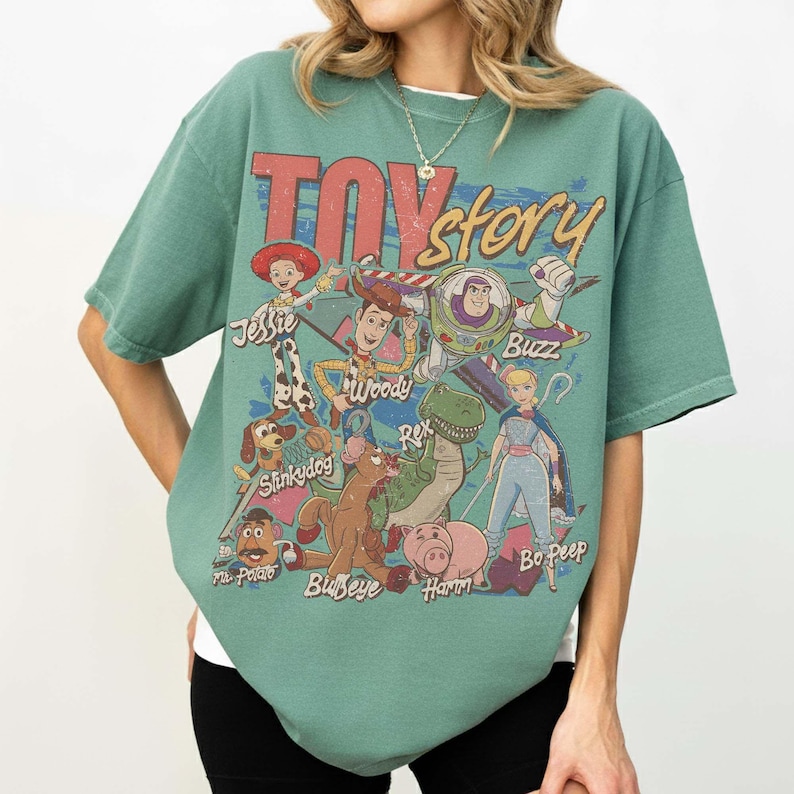 May include: A sage green short-sleeve t-shirt featuring a distressed graphic print of Toy Story characters including Jessie, Woody, Buzz Lightyear, Slinky Dog, Rex, Bullseye, Mr. Potato Head, Hamm, and Bo Peep with the movie title text.