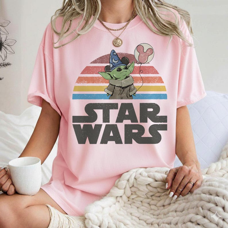 May include: Pink t-shirt featuring a graphic of Grogu from Star Wars wearing a wizard hat and holding a Mickey Mouse balloon. The design includes a retro sunset and the Star Wars logo in gray.