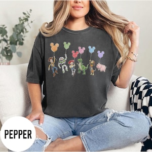 K&ouml;nnte beinhalten: Dunkelgraues T-Shirt mit einer farbenfrohen Grafik von Toy Story-Charakteren, die Mickey Mouse-f&ouml;rmige Ballons halten. Das Wort "PEPPER" ist unten in Wei&szlig; aufgedruckt. Das T-Shirt wird von einer Person getragen.