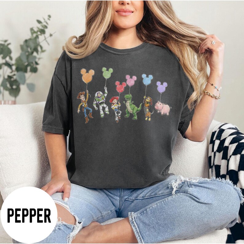 K&ouml;nnte beinhalten: Dunkelgraues T-Shirt mit einer farbenfrohen Grafik von Toy Story-Charakteren, die Mickey Mouse-f&ouml;rmige Ballons halten. Das Wort "PEPPER" ist unten in Wei&szlig; aufgedruckt. Das T-Shirt wird von einer Person getragen.