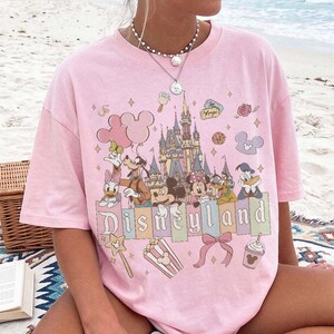 May include: A light pink short-sleeved t-shirt featuring a colourful graphic print of Disneyland with Mickey Mouse, Minnie Mouse, Goofy, Donald Duck, Daisy Duck, and Pluto. The design includes a castle, balloons, popcorn, and a bow.