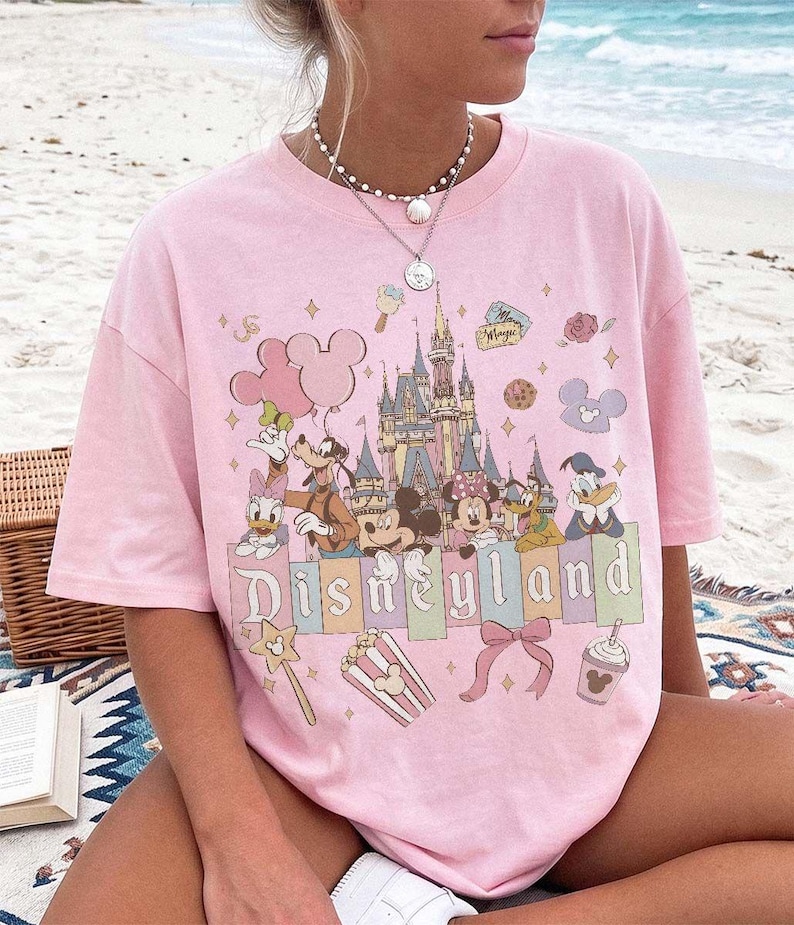 May include: A light pink short-sleeved t-shirt featuring a colourful graphic print of Disneyland with Mickey Mouse, Minnie Mouse, Goofy, Donald Duck, Daisy Duck, and Pluto. The design includes a castle, balloons, popcorn, and a bow.