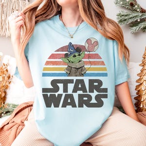 May include: Light blue t-shirt featuring a graphic of Grogu from Star Wars wearing a wizard hat and holding a Mickey Mouse balloon. The design is set against a retro sunset and the Star Wars logo.