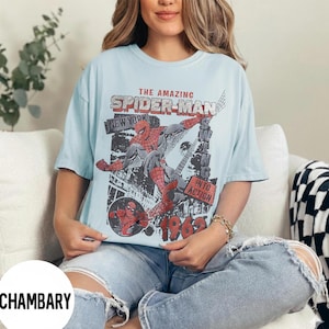 Pu&ograve; includere: T-shirt azzurra con grafica vintage a fumetti di Spider-Man. Il design include il testo "THE AMAZING SPIDER-MAN NEW YORK" e "INTO ACTION 1962". La grafica &egrave; in rosso, nero e grigio.