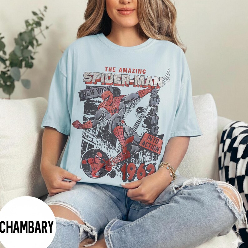 Pu&ograve; includere: T-shirt azzurra con grafica vintage a fumetti di Spider-Man. Il design include il testo "THE AMAZING SPIDER-MAN NEW YORK" e "INTO ACTION 1962". La grafica &egrave; in rosso, nero e grigio.