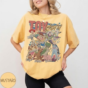 May include: A mustard yellow short-sleeve t-shirt featuring a vintage-style graphic print of Toy Story characters including Woody, Buzz Lightyear, Jessie, Rex, Slinky Dog, Mr. Potato Head, Bullseye, Hamm, and Bo Peep with the text Toy Story.