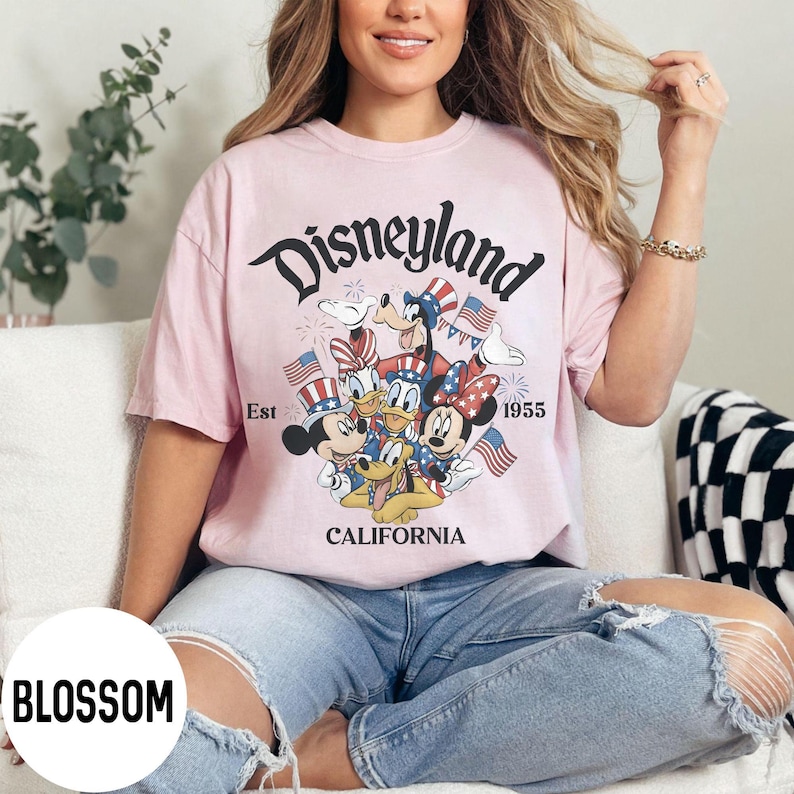 May include: A light pink t-shirt featuring a Disneyland graphic with Mickey Mouse, Donald Duck, Goofy, and Minnie Mouse in patriotic attire. The graphic includes the text "Disneyland California Est 1955".