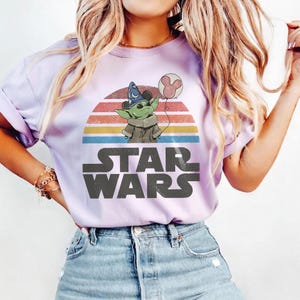 May include: A light purple t-shirt featuring a graphic of Grogu from Star Wars holding a Mickey Mouse balloon. Grogu is wearing a wizard hat. The Star Wars logo is printed below the graphic.