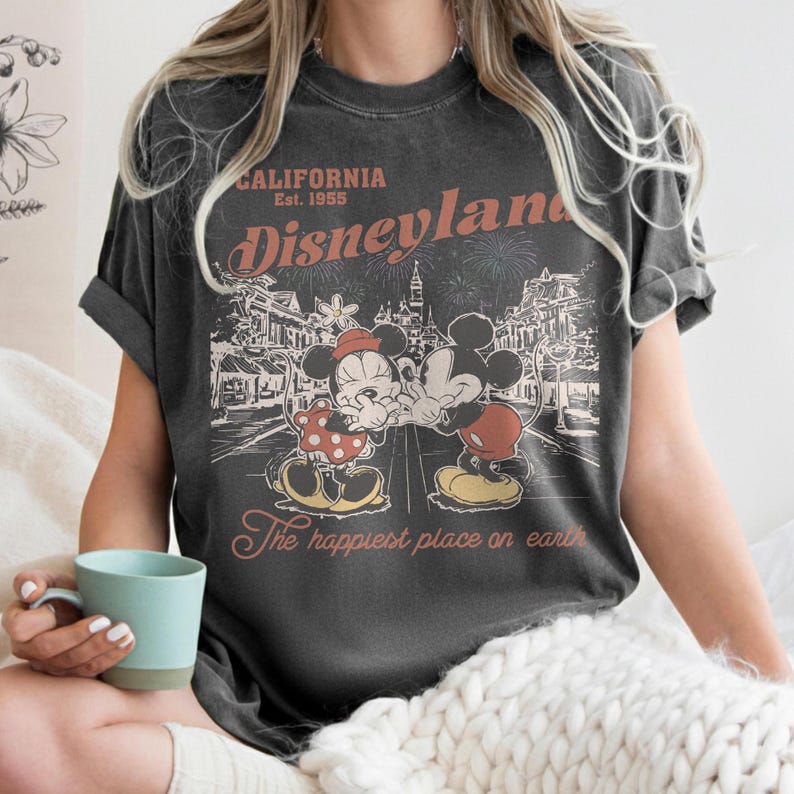 May include: Dark gray t-shirt featuring a vintage Disneyland design. The graphic includes Mickey and Minnie Mouse, fireworks, and the text "CALIFORNIA Est. 1955 Disneyland" and "The happiest place on earth."