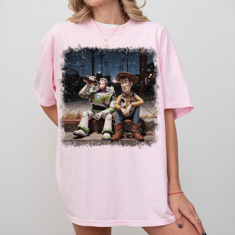 May include: A light pink short-sleeve t-shirt featuring a graphic print of two toy figures sitting on a curb at night. One figure is a white, green, and purple space ranger drinking from a bottle, and the other is a cowboy in a brown hat and vest.