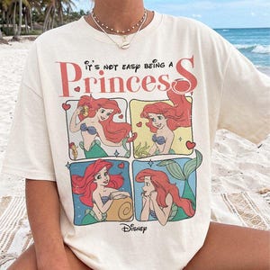 Puede incluir: Una camiseta de manga corta color hueso con un estampado gr&aacute;fico de Ariel de La Sirenita en cuatro paneles distintos. El texto dice "It's not easy being a Princess" en letras rojas y negras, con un peque&ntilde;o logotipo de Disney en la parte inferior.