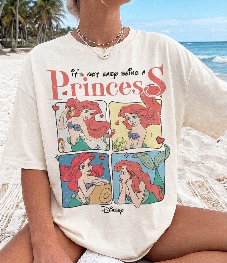 Puede incluir: Una camiseta de manga corta color hueso con un estampado gr&aacute;fico de Ariel de La Sirenita en cuatro paneles distintos. El texto dice "It's not easy being a Princess" en letras rojas y negras, con un peque&ntilde;o logotipo de Disney en la parte inferior.