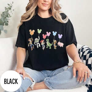 K&ouml;nnte beinhalten: Schwarzes T-Shirt mit einer Grafik von Zeichentrickfiguren, die Mickey Mouse-f&ouml;rmige Ballons halten. Zu den Figuren geh&ouml;ren Woody, Buzz Lightyear, Jessie, Rex, Slinky Dog und Hamm. Das Wort "BLACK" ist unten links aufgedruckt.