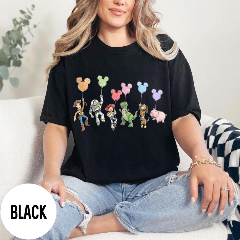 K&ouml;nnte beinhalten: Schwarzes T-Shirt mit einer Grafik von Zeichentrickfiguren, die Mickey Mouse-f&ouml;rmige Ballons halten. Zu den Figuren geh&ouml;ren Woody, Buzz Lightyear, Jessie, Rex, Slinky Dog und Hamm. Das Wort "BLACK" ist unten links aufgedruckt.