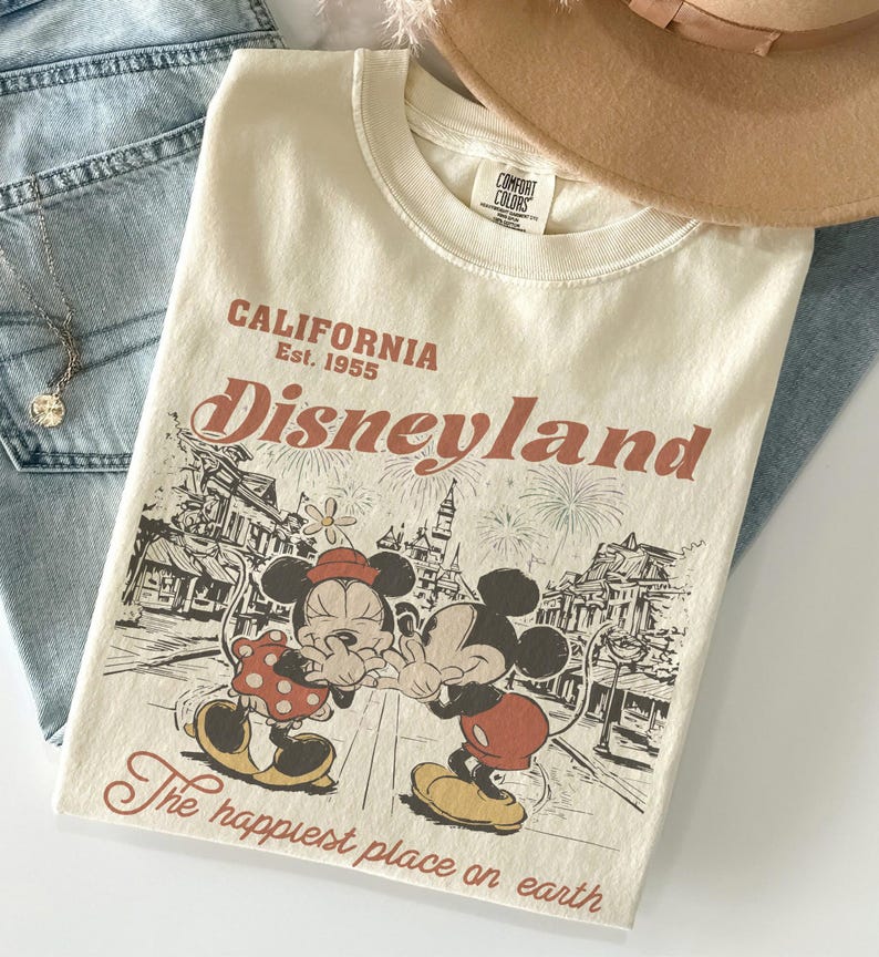 May include: Cream-colored t-shirt with a Disneyland graphic featuring Mickey and Minnie Mouse. The shirt has the text "CALIFORNIA Disneyland Est. 1955" and "The happiest place on earth". The shirt is folded on top of blue jeans.