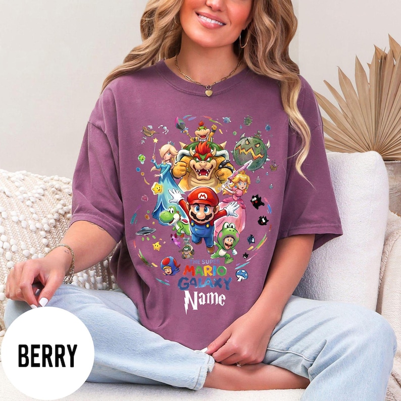 Puede incluir: Camiseta de color bayas con un gr&aacute;fico colorido de Mario y otros personajes del juego Super Mario Galaxy. El dise&ntilde;o incluye el texto "THE SUPER MARIO GALAXY Name". La camiseta es de corte holgado.