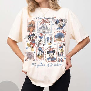 Może przedstawiać: Kremowy t-shirt z kr&oacute;tkim rękawem z nadrukiem graficznym zawierającym napis Walt Disney World, zamek oraz r&oacute;żne postacie, takie jak Myszka Miki, Minnie, Kaczor Donald i Goofy w patriotycznych strojach, z napisem 2026 250 years of freedom.