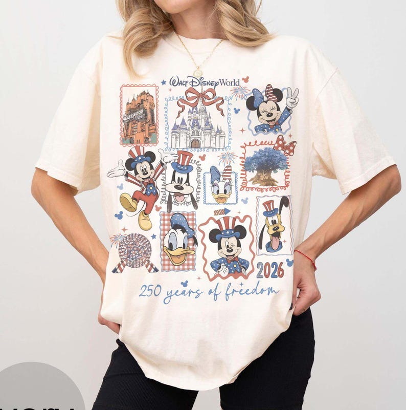 Może przedstawiać: Kremowy t-shirt z kr&oacute;tkim rękawem z nadrukiem graficznym zawierającym napis Walt Disney World, zamek oraz r&oacute;żne postacie, takie jak Myszka Miki, Minnie, Kaczor Donald i Goofy w patriotycznych strojach, z napisem 2026 250 years of freedom.