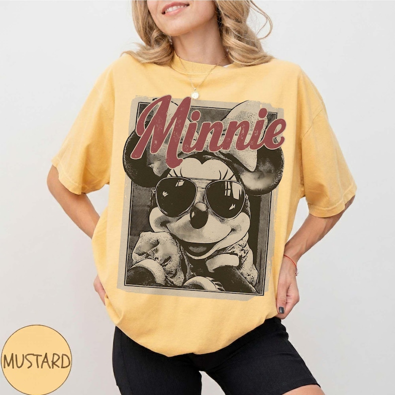 May include: A mustard yellow short-sleeve t-shirt featuring a graphic print of a character wearing sunglasses and a large bow. The text "Minnie" is printed in a dark red script font above the character. The shirt has a crew neckline and a loose fit.