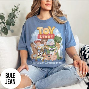K&ouml;nnte beinhalten: Hellblaues T-Shirt mit einer farbenfrohen Grafik von Charakteren aus dem Film "Toy Story". Das Design umfasst Woody, Buzz Lightyear und andere Charaktere, mit den Worten "TOY STORY" und "YOU'VE GOT A FRIEND IN ME" auf dem Shirt.