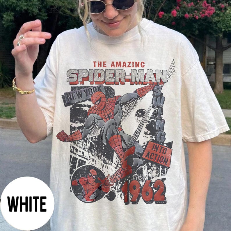 Pu&ograve; includere: T-shirt bianca con grafica di Spider-Man in azione, con il testo "THE AMAZING SPIDER-MAN" e "NEW YORK" in stile fumetto vintage. Viene visualizzato anche l'anno "1962". &Egrave; un capo di abbigliamento casual.