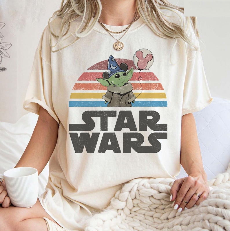 May include: Cream-colored t-shirt featuring a graphic of a green alien character wearing a wizard hat and holding a Mickey Mouse balloon. The design is set against a retro sunset with the words "STAR WARS" in bold, gray letters.
