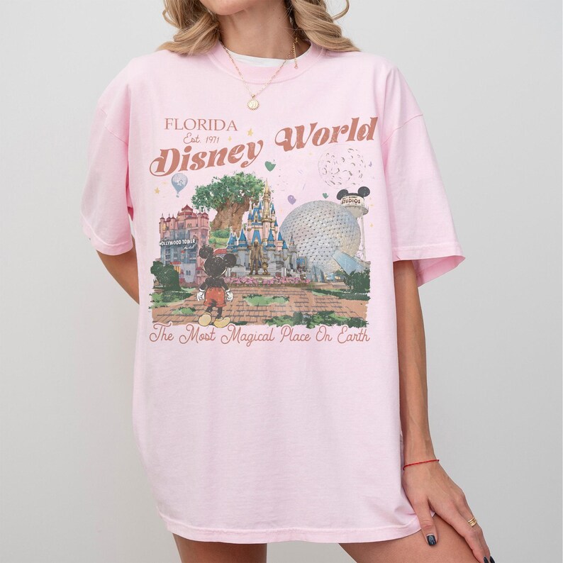 May include: A light pink t-shirt featuring a graphic of Disney World landmarks, including Cinderella's castle and Spaceship Earth. The text "Florida Disney World" and "The Most Magical Place On Earth" are printed on the shirt. The shirt has short sleeves.